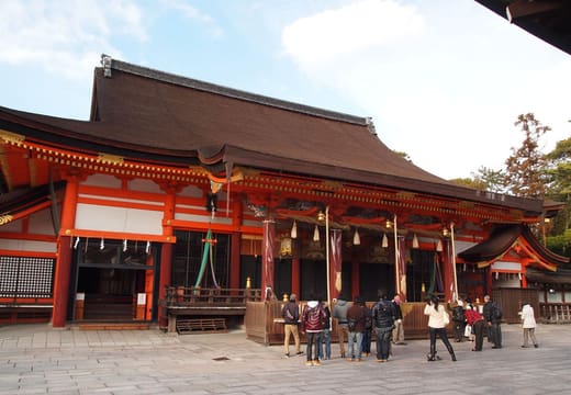 The vermilion main hall of Yasaka Shrine with visitors in its paved courtyard, Kyoto