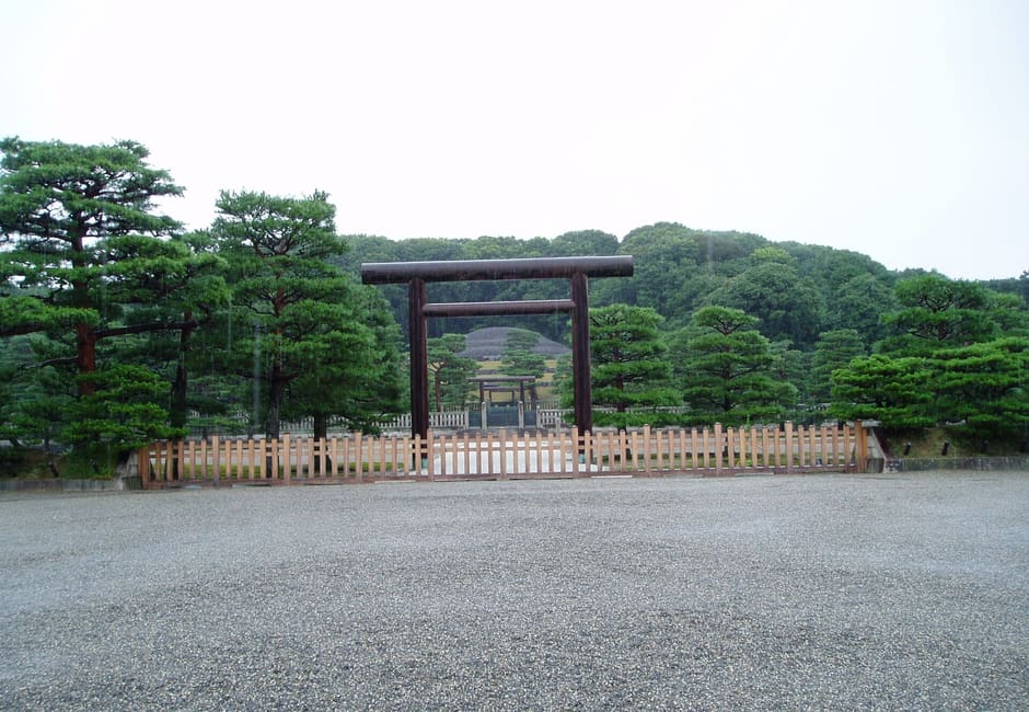 The plain torii and wooden fence at Fushimi Momoyama Mausoleum, with Emperor Meiji's stone-covered tomb mound beyond in the trees