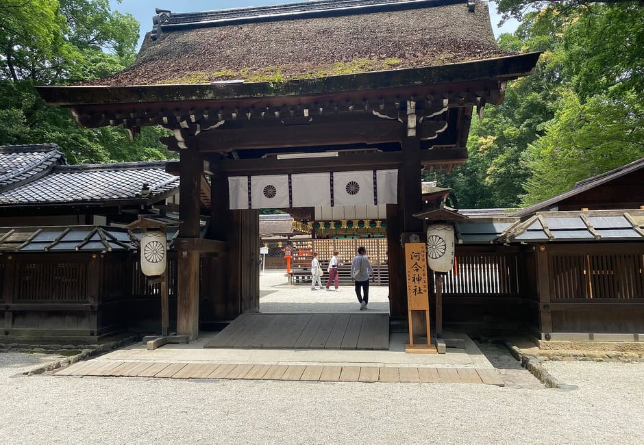 The gate of Kawai Shrine with its moss-covered roof, the shrine name board and a round paper lantern on each side