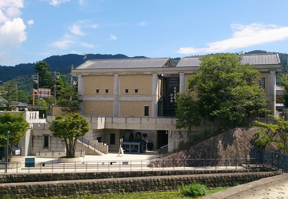 The Lake Biwa Canal Museum building with its name on the front, stone canal walls below it and the Higashiyama hills behind