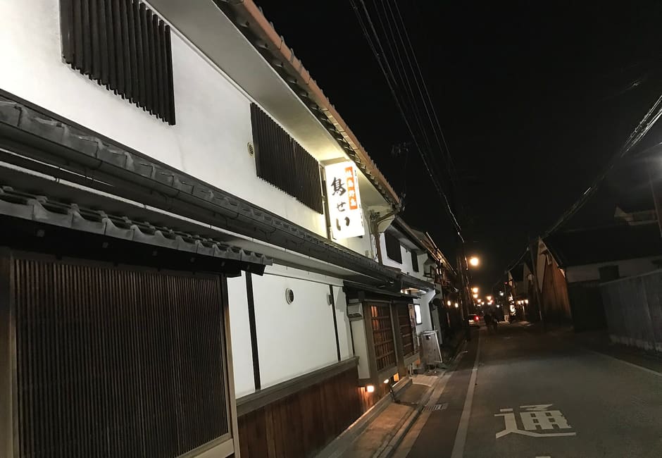 The white sake storehouse of Torisei Honten in Fushimi at night, its lit sign hanging over the street