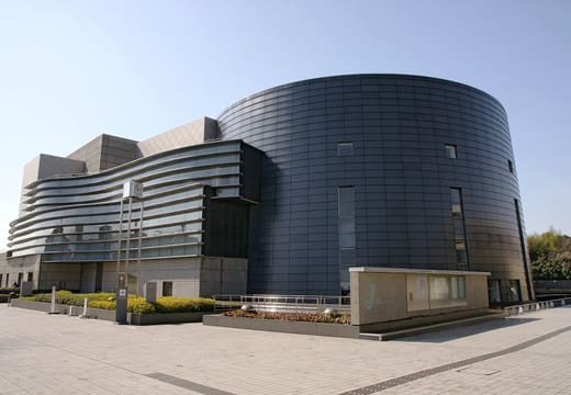 The curved dark metal wall of Kyoto Concert Hall in Kitayama beside a paved plaza